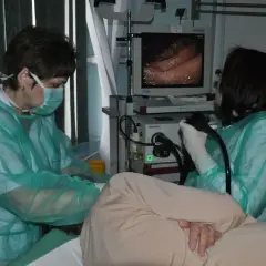 Lekarze w zielonych fartuchach przeprowadzają gastroskopię, mimo że jeden z nich ma przeziębienie. Obraz z endoskopu pokazuje wnętrze żołądka.