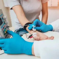 Lekarz w niebieskich rękawiczkach wykonuje próbę tężyczkową, przykładając elektrody do dłoni pacjentki i obsługując aparat.