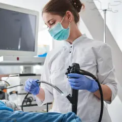 Lekarka w masce i rękawiczkach przygotowuje się do gastroskopii. Pacjent leży, a na ekranie widać obraz z kamery. Po zabiegu może pojawić się ból po gastroskopii.