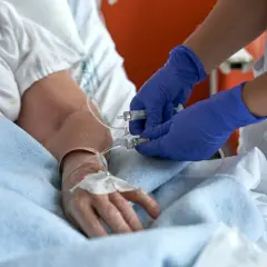 Pielęgniarka w niebieskich rękawiczkach przygotowuje się do usuwania cewnika w domu pacjenta, podłączając strzykawkę do linii infuzyjnej.