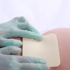 Ręce w rękawiczkach przykładają kwadratowy plaster na odleżyny na kości ogonowej.