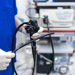 Lekarz w białych rękawiczkach trzyma endoskop, przygotowując się do gastroskopii. Sprzęt medyczny w tle.