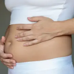 Kobieta dotyka swojego wzdętego brzucha, ubranego w biały top i majtki.