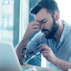 Mężczyzna z zarostem siedzi przy biurku, masując skronie. W ręku trzyma okulary, a przed nim leży laptop. Widać jego złe samopoczucie.