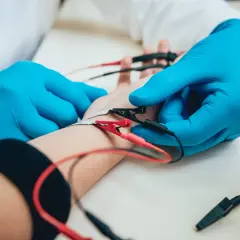 Lekarz w niebieskich rękawiczkach przeprowadza badanie EMG, podłączając elektrody do przedramienia pacjenta.