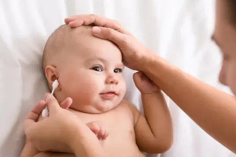 Delikatne czyszczenie uszu niemowlaka. Mama ostrożnie używa patyczka higienicznego, by zadbać o higienę maluszka.