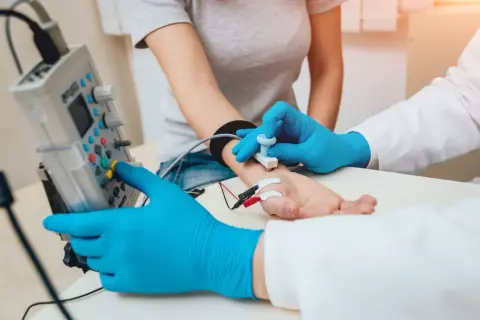 Lekarz w niebieskich rękawiczkach wykonuje próbę tężyczkową, przykładając elektrody do dłoni pacjentki i obsługując aparat.