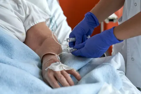 Pielęgniarka w niebieskich rękawiczkach przygotowuje się do usuwania cewnika w domu pacjenta, podłączając strzykawkę do linii infuzyjnej.