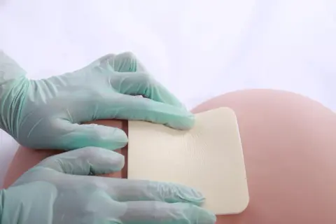 Ręce w rękawiczkach przykładają kwadratowy plaster na odleżyny na kości ogonowej.