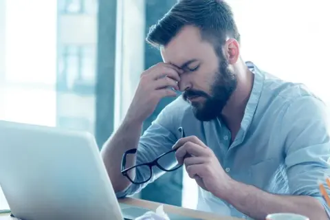 Mężczyzna z zarostem siedzi przy biurku, masując skronie. W ręku trzyma okulary, a przed nim leży laptop. Widać jego złe samopoczucie.