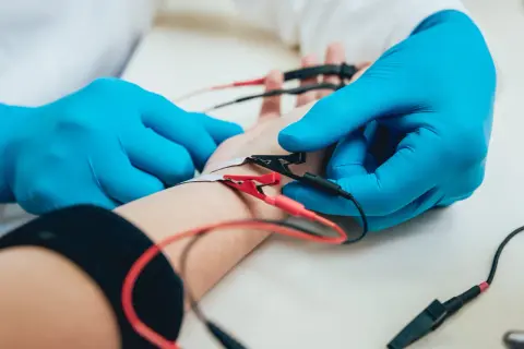 Lekarz w niebieskich rękawiczkach przeprowadza badanie EMG, podłączając elektrody do przedramienia pacjenta.