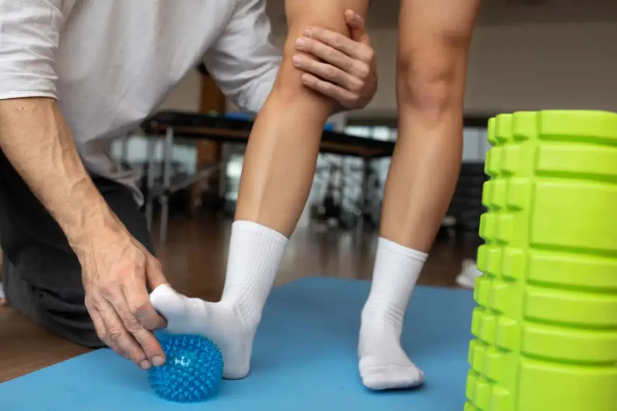 Fizjoterapeuta masuje stopę pacjenta niebieską piłeczką, co może być pomocne co na skręcenie kostki. Obok zielony wałek do masażu.