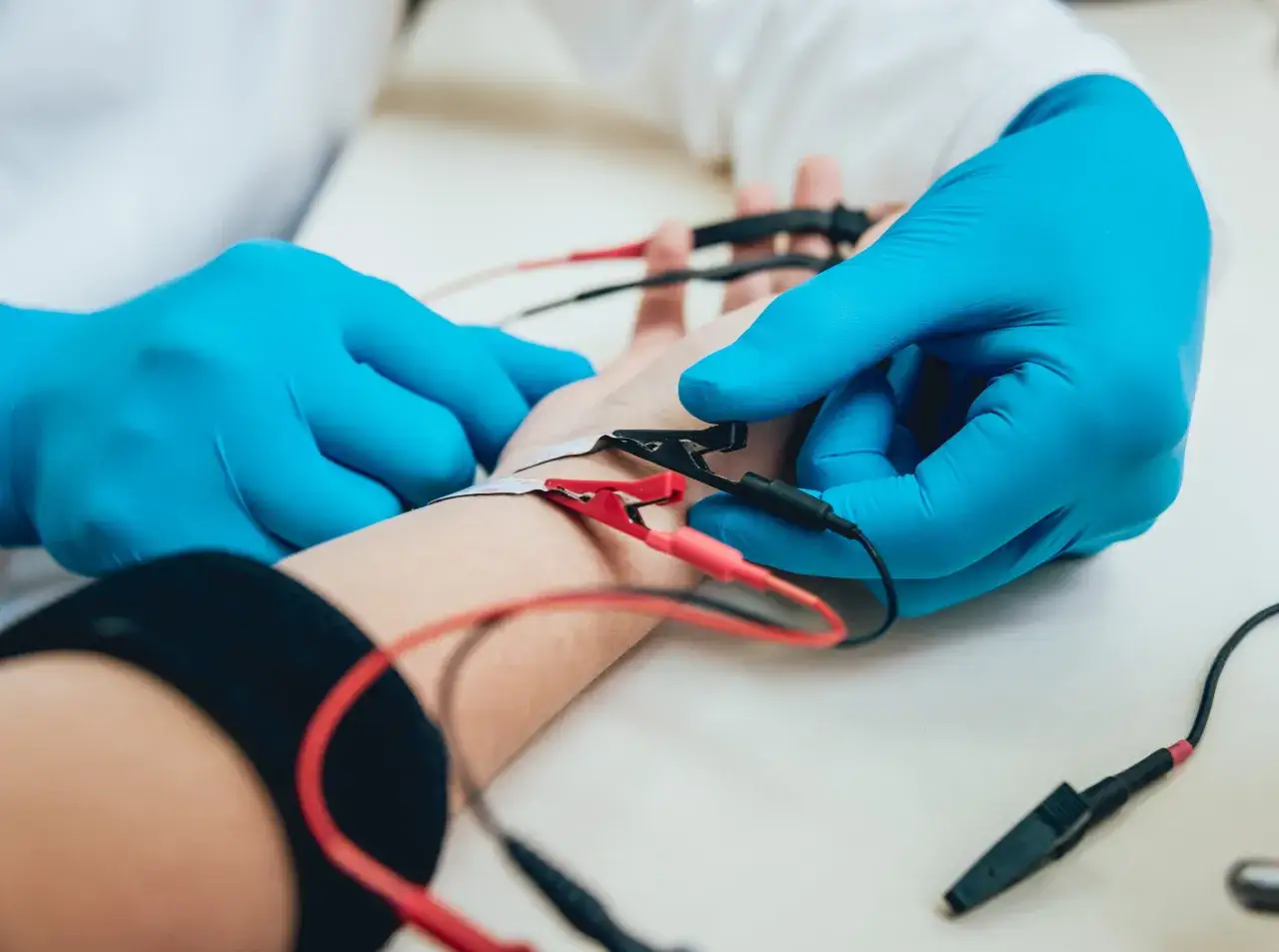Lekarz w niebieskich rękawiczkach wykonuje próbę tężyczkową, podłączając elektrody do przedramienia pacjenta.