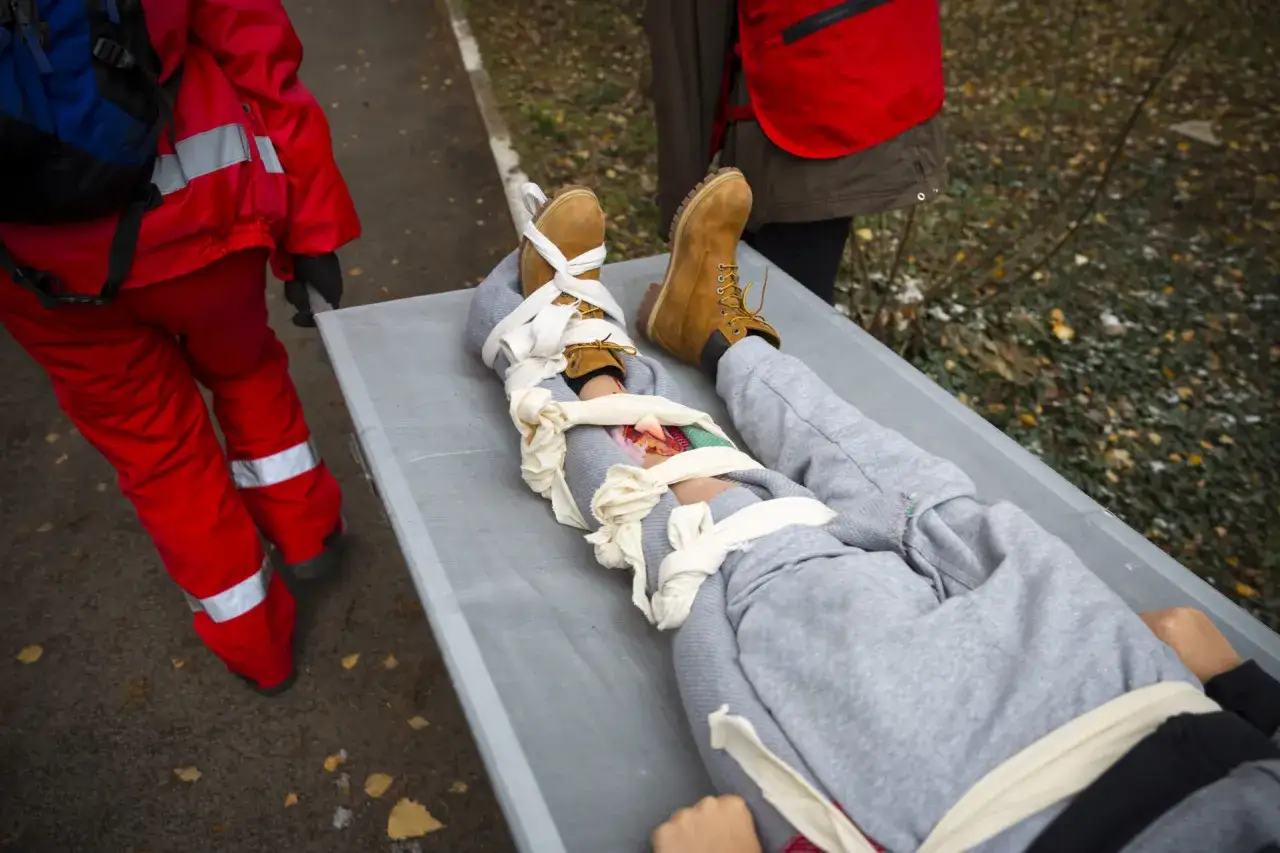 Na noszach leży osoba z widocznym złamaniem otwartym nogi, opatrzonym bandażami. Ratownik w czerwonym stroju zabezpiecza ranę.